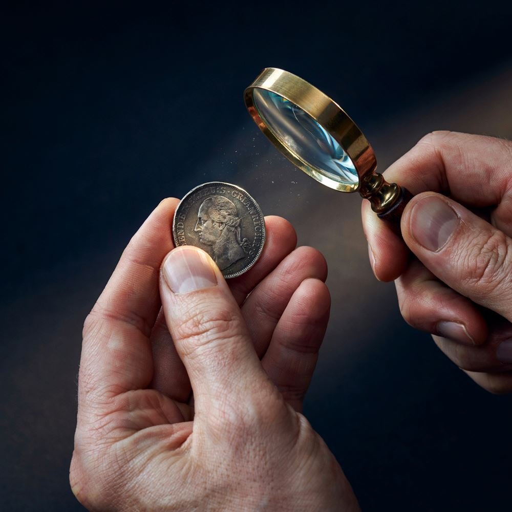 Mãos de numismata examinando uma moeda de prata com lupa